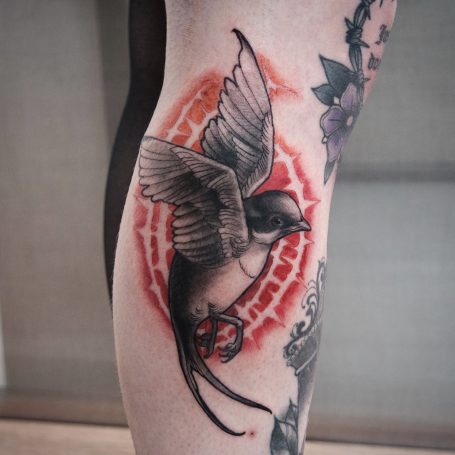 Tattoo eines fliegenden Vogels mit rotem Hintergrund auf dem Oberschenkel.